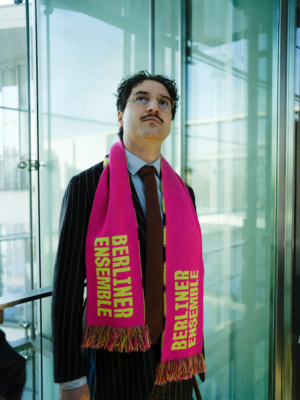 Patrick Pehl in einem dunkelbraunen Nadelstreifenanzug und brauner Krawatte trägt einen neon-rosanen Berliner-Ensemble-Fanschal