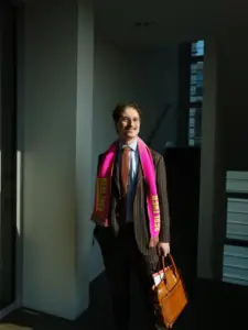 Patrick Pehl mit dem neonrosanen Fanschal „Drama“ des Berliner Ensemble (BE) im Bundestag in einem modernen Treppenhaus und einer Aktentasche. Patrick Pehl lächelt und trägt einen gestreiften Anzug in schokoladenbraun mit einer brauen Krawatte.